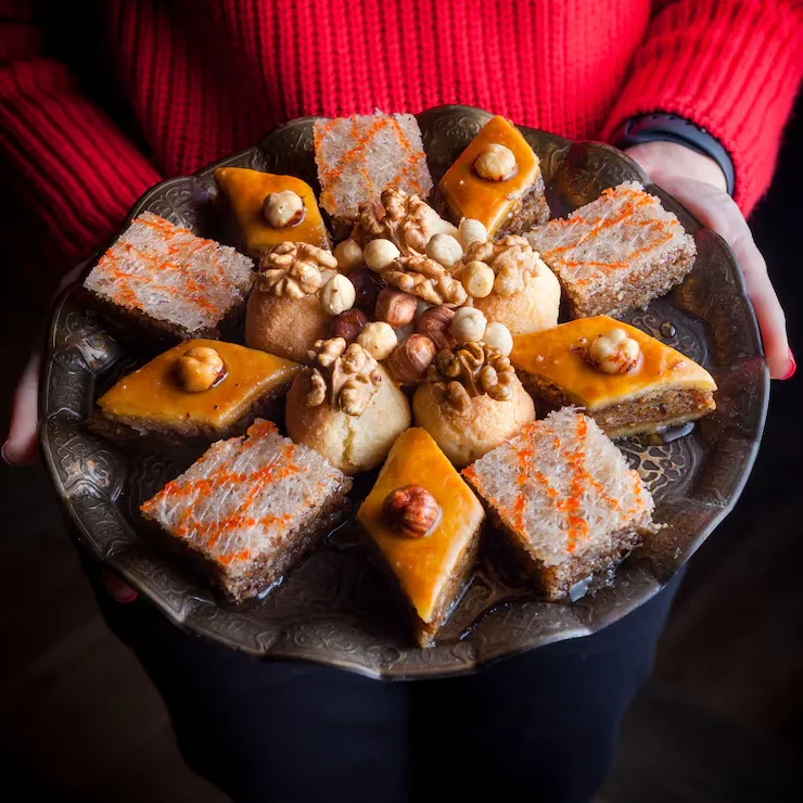 Hands presenting ornate brass platter with Afghan sweets including baklava, halwa, and nut-topped desserts