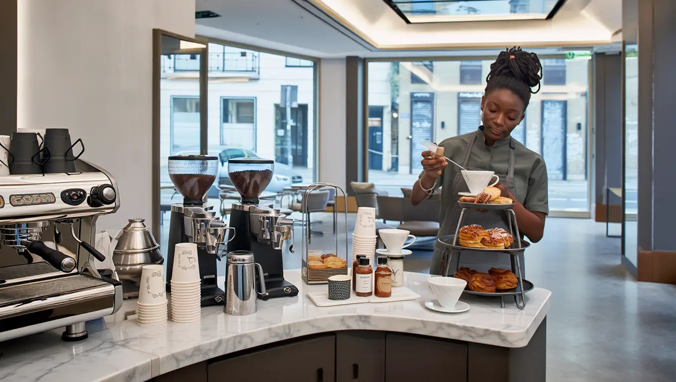 Professional barista arranging fresh pastries on tiered stand beside commercial espresso machine in modern office