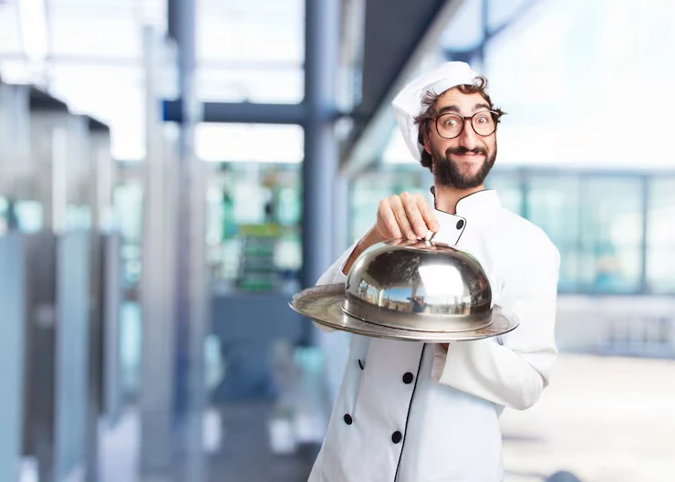 Smiling chef in white uniform presenting covered dish with silver cloche in modern kitchen