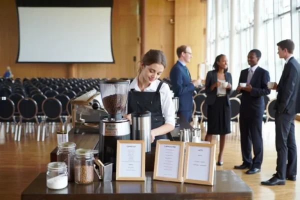 Barista preparing coffee at a conference venue with a mobile coffee station while attendees network during a seminar break.