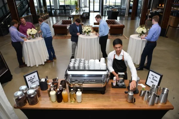 Barista preparing drinks at a custom coffee station during a social event with guests networking around decorated cocktail tables.