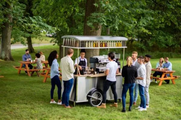 Barista serving drinks from a mobile coffee bar at an outdoor event while guests gather around and others relax at picnic tables in a park setting.