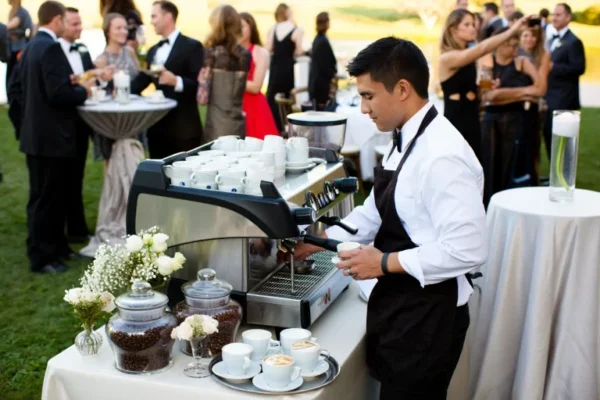 Barista preparing coffee at an outdoor wedding reception with decorative coffee setups and elegantly dressed guests in the background.