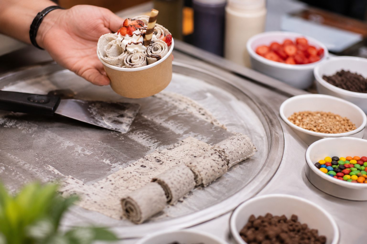 Hand holding a customised rolled ice cream dessert with assorted toppings at a professional dessert bar station, surrounded by fresh ingredients and interactive live catering setup.