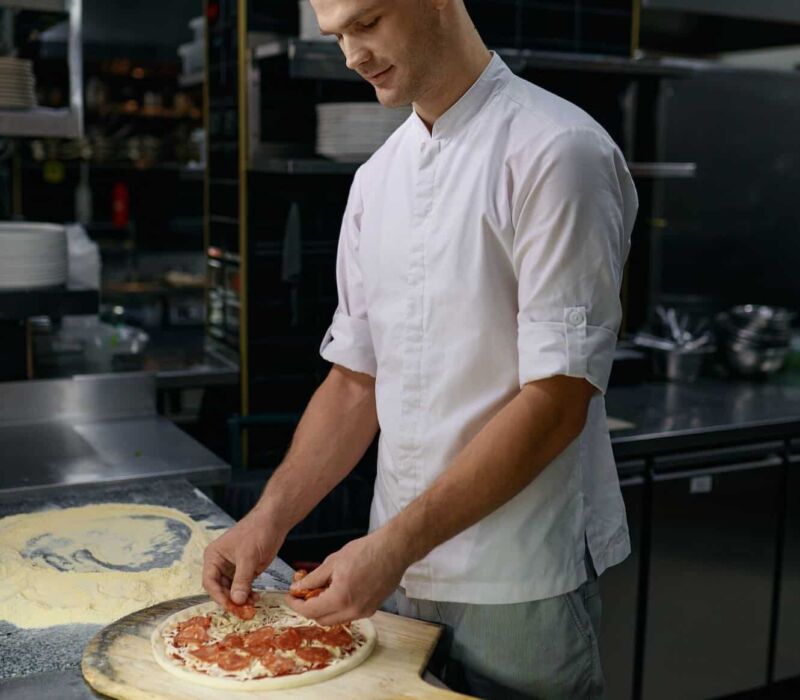 Pizzaiolo adding ham on pizza base with cheese, preparing italian dish